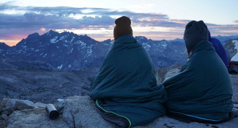 Two people wrapped in blankets sit on a rock, overlooking the vast mountain area below. 